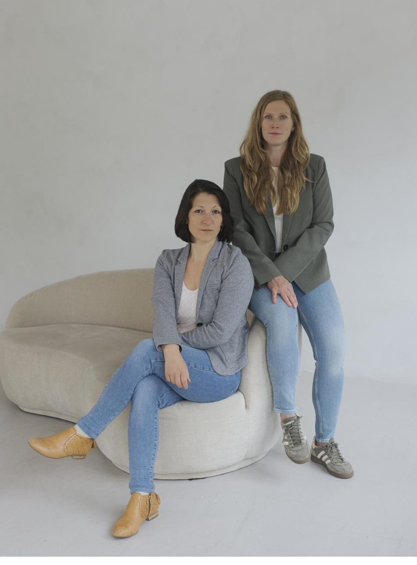 Julia Menzler sitzt auf einem Sofa, Stefanie Bayer lehnt daneben